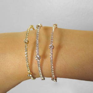 May include: Three delicate bracelets, each with a different metal finish: gold, silver, and rose gold. Each bracelet features a line of small, sparkling stones and a larger, central stone. The bracelets are displayed on a wrist.