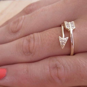 May include: A gold-colored arrow ring with a unique design. The ring features an arrow pointing down and another arrow pointing up, both adorned with small, clear gemstones. The ring is worn on a finger, showcasing its delicate details and craftsmanship.
