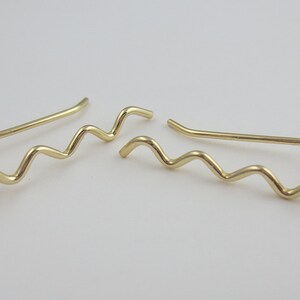 May include: A pair of gold-colored ear climbers. Each climber features a wavy design that curves up the ear. The earrings are made of a shiny, metallic material and are displayed against a white background.