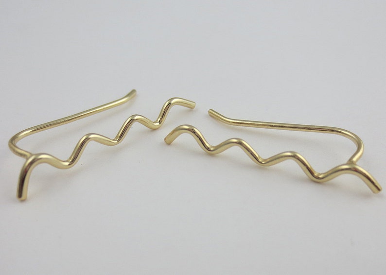 May include: A pair of gold-colored ear climbers. Each climber features a wavy design that curves up the ear. The earrings are made of a shiny, metallic material and are displayed against a white background.