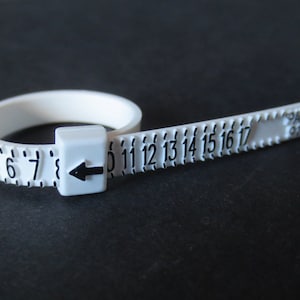 May include: A white plastic ring sizer with black numbers from 5 to 17. An arrow indicates the size. The text "PAT PEND 9101693" is printed on the band. The tool is used to measure ring sizes.
