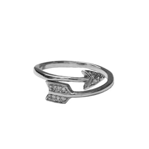 May include: A silver-colored ring in the shape of an arrow. The arrow design features small, clear gemstones. The ring wraps around the finger, with the arrowheads pointing in opposite directions. The ring is set against a white background.