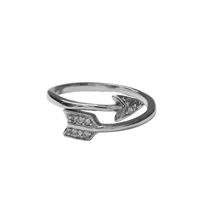 May include: A silver-colored ring in the shape of an arrow. The arrow design features small, clear gemstones. The ring wraps around the finger, with the arrowheads pointing in opposite directions. The ring is set against a white background.