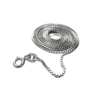 May include: A silver-colored box chain necklace with a lobster clasp, coiled on a white background. The chain is made of small, square links, creating a textured appearance. The clasp is open, showing its mechanism.