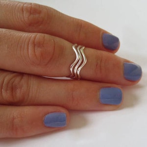 May include: A silver-colored ring with a wavy design is worn on a finger. The ring has three stacked, undulating bands. The nails are painted a light blue color. The background is white.