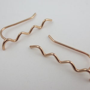 May include: A pair of rose gold ear climbers. Each earring features a wavy design that curves up the ear. The earrings have a simple, elegant style. The metal has a warm, pinkish hue.