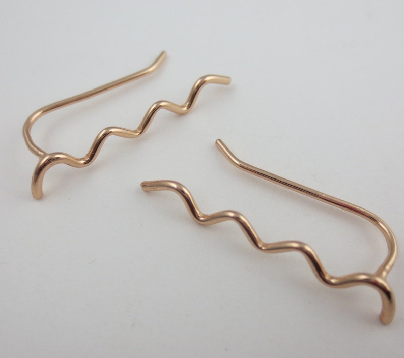 May include: A pair of rose gold ear climbers. Each earring features a wavy design that curves up the ear. The earrings have a simple, elegant style. The metal has a warm, pinkish hue.