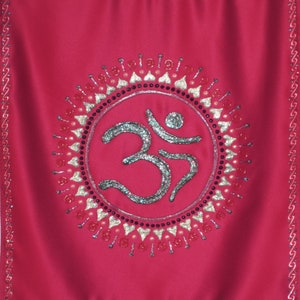 May include: A red fabric wall hanging with a large, glittery Om symbol surrounded by a circle of red, gold, and white beads.