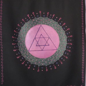 Könnte beinhalten: Ein schwarzer Wandbehang aus Stoff mit einem geometrischen Muster in Rosa und Lila. Das Muster zeigt einen Kreis mit einem Davidstern in einem Dreieck. Der Kreis ist von einem grünen und silbernen Glitzer-Mandala umgeben.