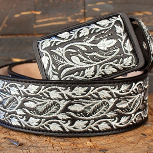May include: A black leather belt with silver embroidery of a floral design. The belt has a silver buckle.