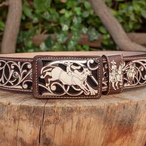 Może przedstawiać: Brązowy skórzany pasek z srebrną klamrą przedstawiającą kowboja na koniu. Pasek ma wycięty wzór z motywami kwiatowymi.