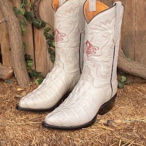 May include: A pair of off-white cowboy boots with red floral embroidery. The boots have a textured pattern and a black sole. The background includes a wooden fence and hay.