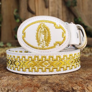 May include: White leather belt with gold embroidered detailing. The belt buckle features an oval design with a gold embroidered image of the Virgin Mary. The belt strap has a geometric pattern in gold embroidery.