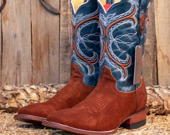 Pode incluir: Um par de botas de cowboy de camurça marrom com cano de couro azul adornado com intrincados pespontos brancos. As botas têm bico fino e salto alto.