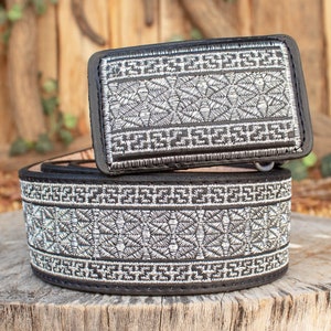 May include: A black leather belt with a silver embroidered design. The belt has a matching buckle with a silver embroidered design.