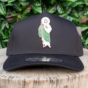 May include: A black baseball cap with a green and white saint figure pin on the front. The cap has a black brim and a black adjustable strap.