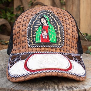 以下が含まれることがあります： 茶色の織り模様のトラッカーハット。黒いメッシュの背面と湾曲したつばが特徴です。フロントパネルには、赤と緑の宗教的な人物の刺繍画像が表示されています。つばは白と茶色のデザインです。