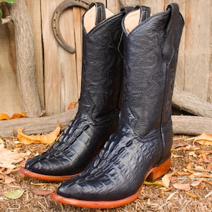 Può includere: Un paio di stivali da cowboy neri con un motivo strutturato a forma di coccodrillo sul piede e sul collo del piede. Gli stivali hanno un gambale alto, una punta a punta e una suola marrone rossiccio. Lo sfondo include una staccionata di legno e un ferro di cavallo.