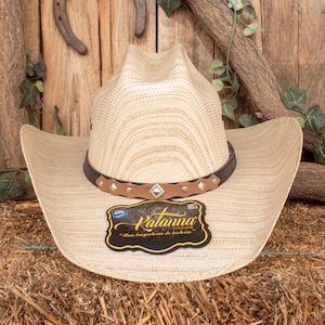 May include: A beige straw cowboy hat with a brown leather band and silver accents. The hat has a woven texture and a wide brim. The hat is sitting on a bale of hay. The hat has a label that says "Kalamna Gold Line" and "Una trayectoria de historia."