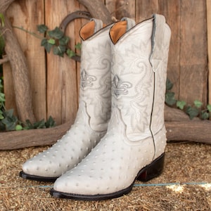 May include: A pair of white ostrich leather cowboy boots with intricate floral embroidery. The boots have a pointed toe and a stacked heel.
