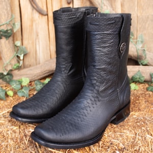 Peut inclure: Une paire de bottes de cowboy en cuir noir avec un motif texturé. Les bottes ont un bout carré, un petit talon et une fermeture éclair sur le côté. Les bottes sont exposées sur un lit de foin.