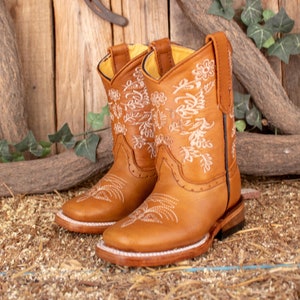 Könnte beinhalten: Ein Paar braune Leder-Cowbo Stiefel mit weißen Blumenstickereien. Die Stiefel haben eine spitze Zehenpartie und einen niedrigen Absatz.