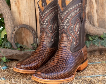Pode incluir: Um par de botas de cowboy de couro castanho com um padrão de pele de cobra texturizado no pé e um design decorativo costurado na parte superior da bota. As botas têm bico quadrado e sola de madeira.