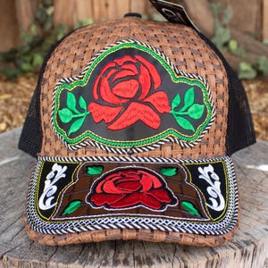 Peut inclure: Casquette de camionneur marron tressée avec un dos en filet noir et une visière incurvée. La casquette présente une grande rose rouge brodée sur un patch noir, encadrée de feuilles vertes et d'une bordure décorative. La visière a aussi une rose.