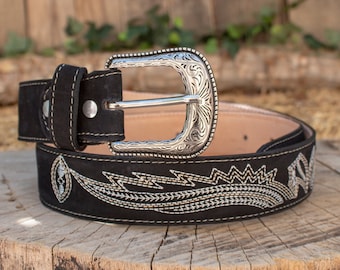 May include: Black suede belt with a silver-coloured, ornate buckle. The belt features detailed white stitching and decorative patterns. The belt is displayed on a wooden surface, showcasing its craftsmanship and design.