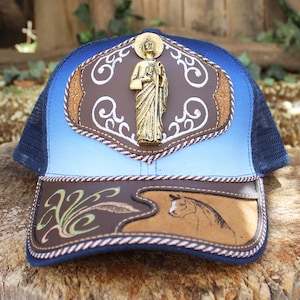 May include: A blue and brown trucker hat with a gradient design. The hat features a gold-colored religious figure on the front, along with brown leather patches and decorative stitching. A horse head is also visible on the brim.
