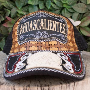 May include: A black and brown trucker hat with the word "AGUASCALIENTES" embroidered on the front. The hat features a woven pattern on the front panel and a mesh back. The brim is black with decorative stitching and a white emblem.