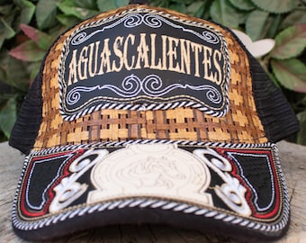 Może przedstawiać: Czarna i brązowa czapka typu trucker z napisem "AGUASCALIENTES" wyhaftowanym z przodu. Czapka ma tkany wzór na przednim panelu i siatkowy tył. Daszek jest czarny z ozdobnymi przeszyciami i białym emblematem.