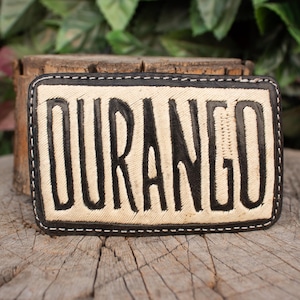 May include: Rectangular leather belt buckle with the word "DURANGO" in bold black letters. The buckle has a cream-colored center with a black border and white stitching. The buckle is likely for a belt.