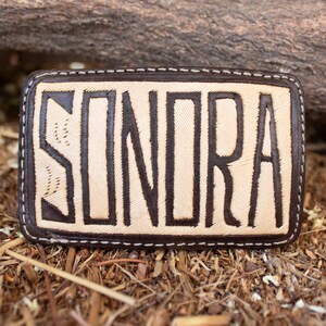 May include: Rectangular leather belt buckle with the word "SONORA" in bold, dark brown letters. The letters are set against a light tan background, with a dark brown border and white stitching. The buckle is resting on a bed of dry leaves and twigs.