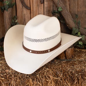 Könnte beinhalten: Ein weißer Cowboyhut mit einem braunen Lederband und silberfarbenen Nieten. Der Hut hat eine belüftete Krone und eine breite Krempe. Der Hut liegt auf einem Heubett.