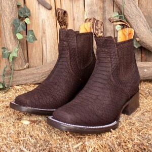 Peut inclure: Une paire de bottines en cuir marron avec un motif texturé. Les bottes ont un bout carré, un petit talon et une tirette avec un logo. Les bottes sont présentées sur un lit de paille devant un fond en bois.