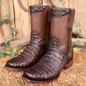 Peut inclure: Une paire de bottes de cowboy en cuir marron avec un motif peau de crocodile sur le pied et la tige. Les bottes ont un bout carré, un dessus décoratif et une fermeture éclair latérale. Les bottes sont exposées sur de la paille.