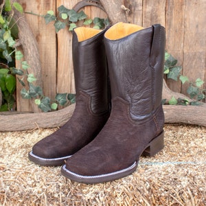 Può includere: Un paio di stivali da cowboy in pelle marrone con finitura in camoscio. Gli stivali hanno una punta a punta e un tacco basso.