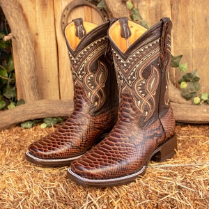 Op de afbeelding: Een paar bruine leren cowboylaarzen met ingewikkelde borduursels en een slangenhuidpatroon. De laarzen hebben een spitse neus en een lage hak.