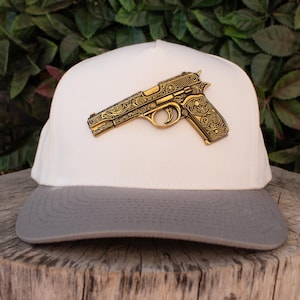 Puede incluir: Una gorra de béisbol blanca y gris con un detallado emblema de pistola dorada. La gorra tiene un diseño clásico de gorra de camionero con un panel frontal blanco y una visera gris. La pistola tiene intrincados adornos.