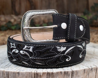 May include: Black belt with a silver buckle and floral embroidery. The belt features a decorative silver buckle with an engraved design. The black leather belt has white stitching and a detailed floral pattern.