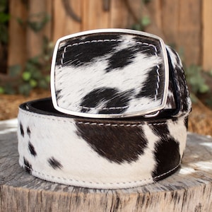 May include: A cowhide belt with a silver-colored rectangular buckle. The belt features a black and white spotted pattern. The buckle is stitched to the belt with white thread. The belt is displayed on a wooden surface.