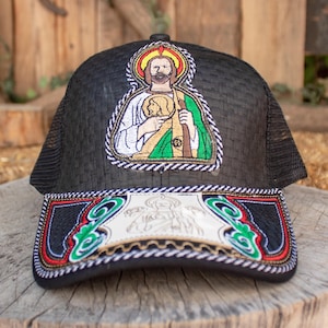 May include: Black trucker hat with a woven fabric crown and a mesh back. The hat features an embroidered image of a saint with a halo and a staff. The brim of the hat has a decorative trim with green, red, and gold accents.