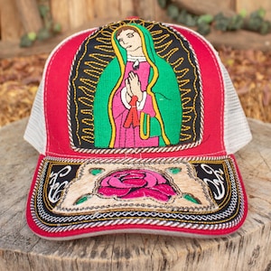 May include: Red trucker hat with a white mesh back and a detailed embroidered design. The design features an image of a religious figure with a green shawl and pink clothing. The brim has a rose and decorative stitching.