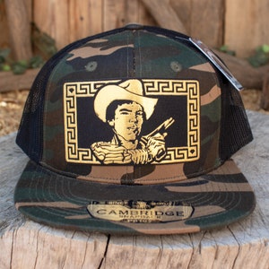 May include: A black and green camouflage trucker hat with a gold embroidered patch featuring a cowboy holding a gun. The patch has a gold Greek key border. The hat has a black mesh back and a gold "CAMBRIDGE SNAPBACK" label.