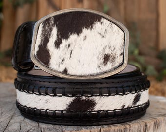 May include: A black and white cowhide belt with a silver-toned buckle. The belt features a section of cowhide with black stitching and a matching cowhide buckle. The belt is resting on a wooden surface.