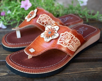 May include: A pair of brown leather sandals with floral cutouts and embossed designs. The sandals have a thong strap and a dark sole. The sandals are decorated with white stitching and a small flower with a green center.