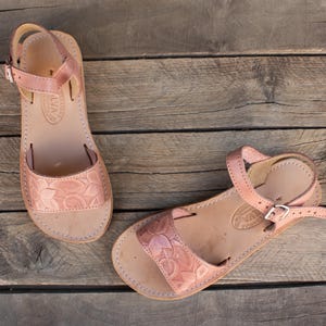May include: Two light brown leather sandals with floral embossed straps and ankle straps. The sandals are displayed on a weathered wooden surface. The sandals have a buckle closure and are designed for comfort and style.