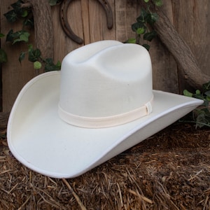 Peut inclure: Un chapeau de cowboy blanc avec un large bord et un ruban crème. Le chapeau est en feutre et repose sur de la paille. Un fer à cheval et des planches de bois sont en arrière-plan.