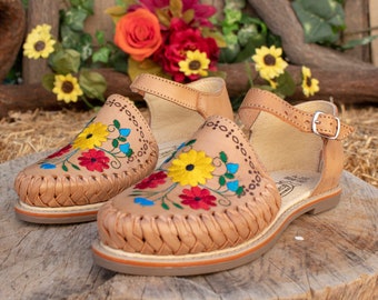 May include: A pair of tan leather sandals with colorful floral embroidery. The sandals have a woven strap and an ankle strap with a buckle.
